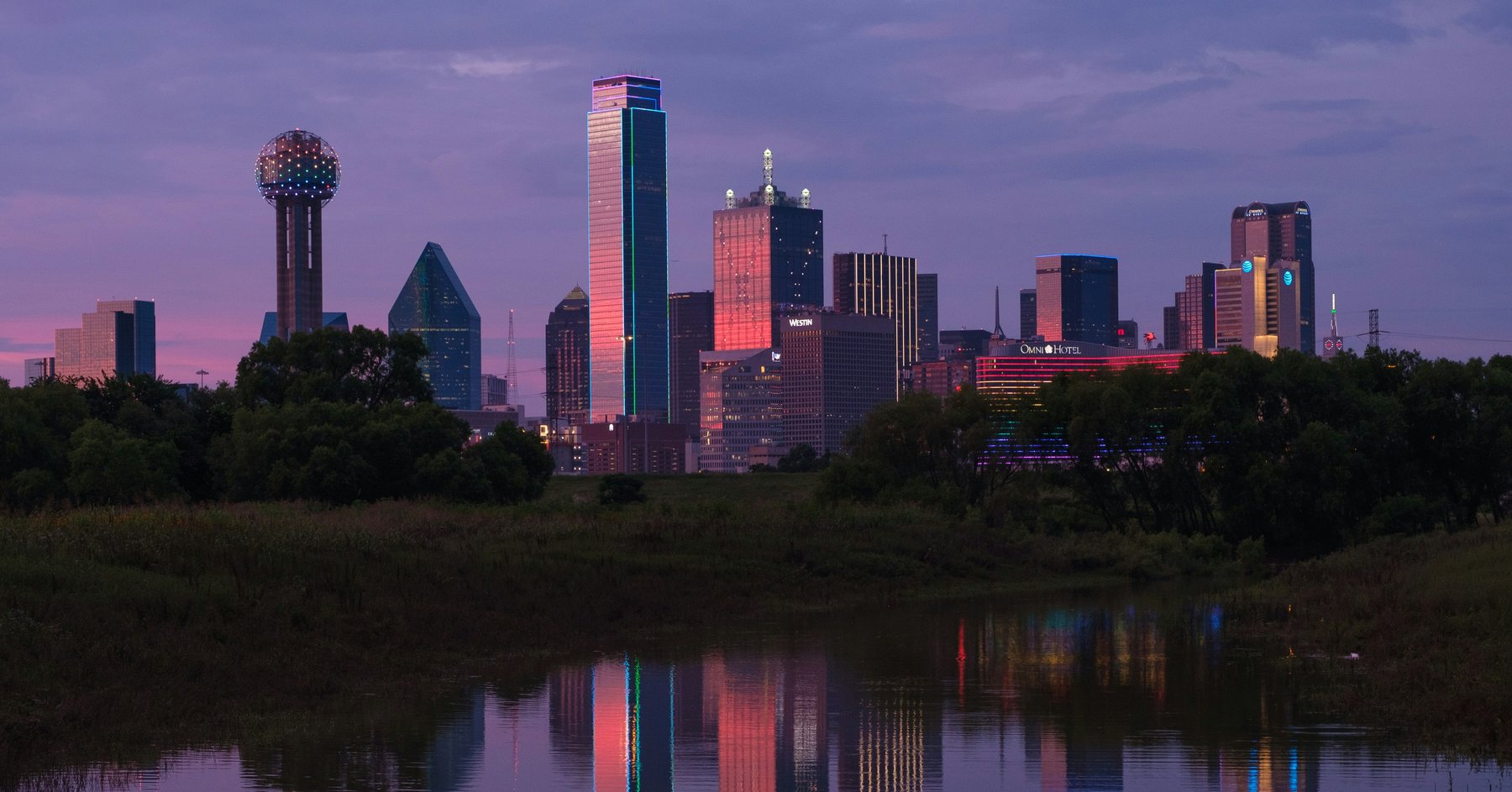 Downtown Dallas skyline at dusk — Reunion Tower, Bank of America Plaza, and the Fountain Place buildings reflected in the Trinity River